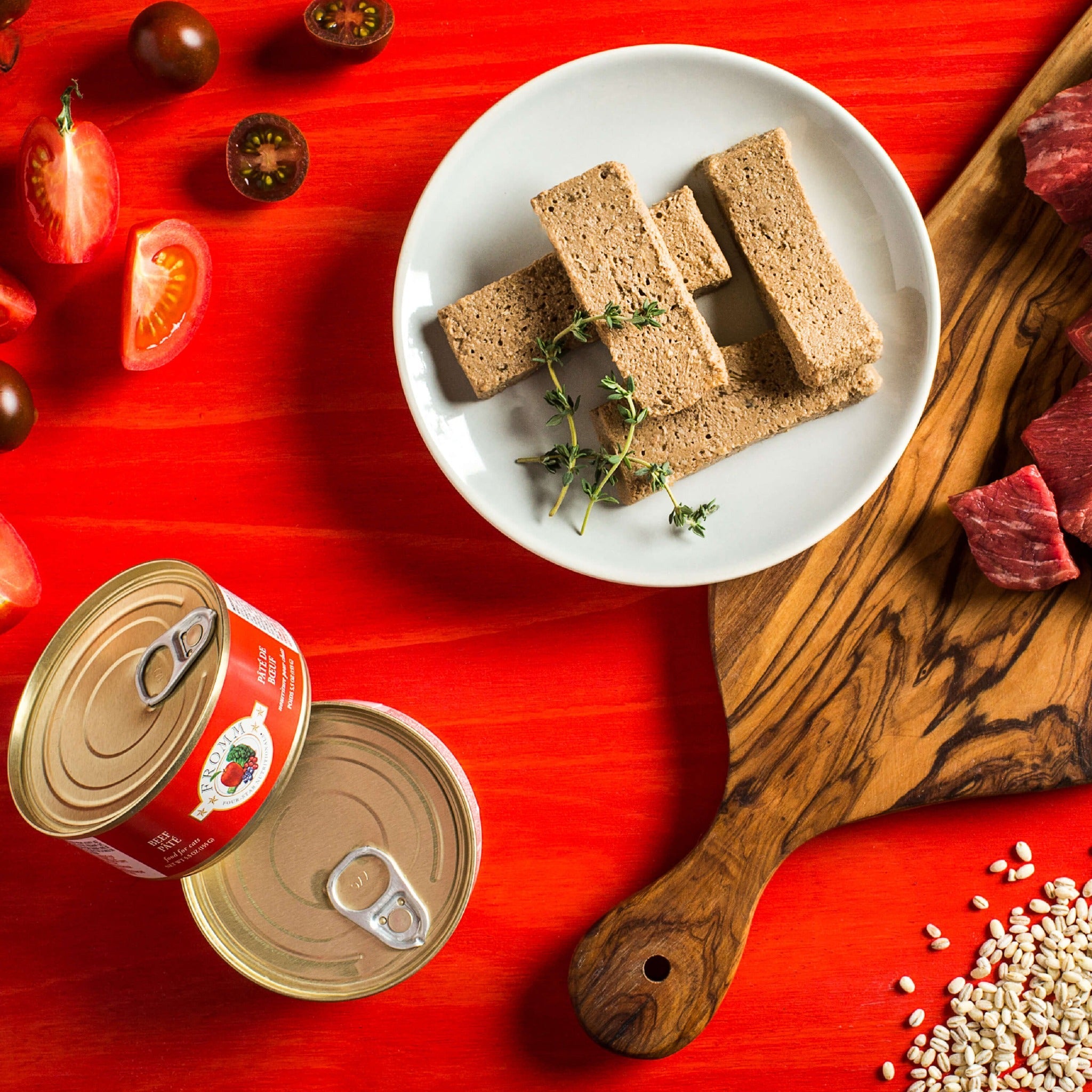 Raw ingredients of Fromm Four-Star Beef Pâté: beef cuts, tomatoes, and barley beside sliced pâté on a plate with two cans