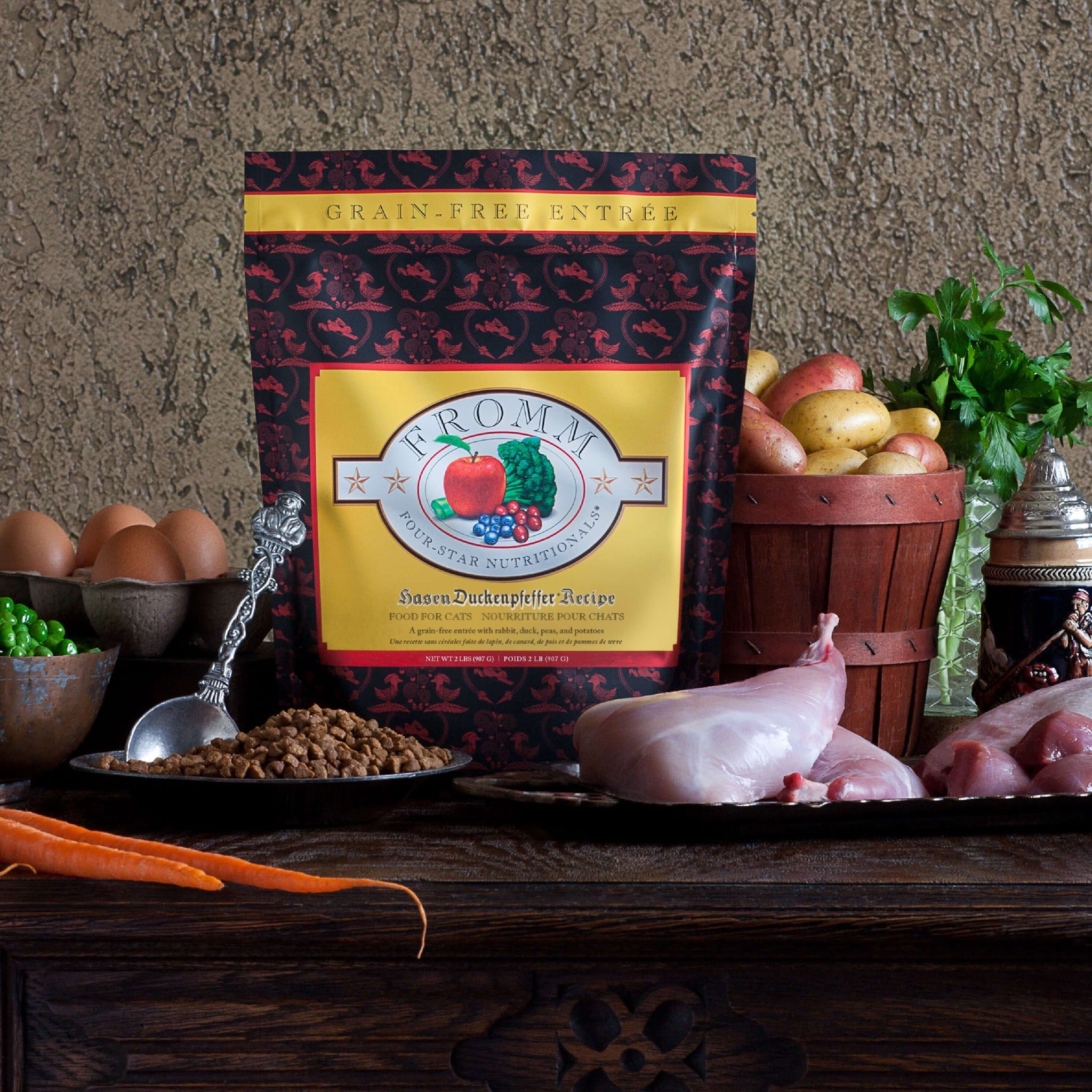 Bag of Fromm Four-Star Hasen Duckenpfeffer cat food on a table with raw rabbit, duck, peas, potatoes, carrots, and eggs