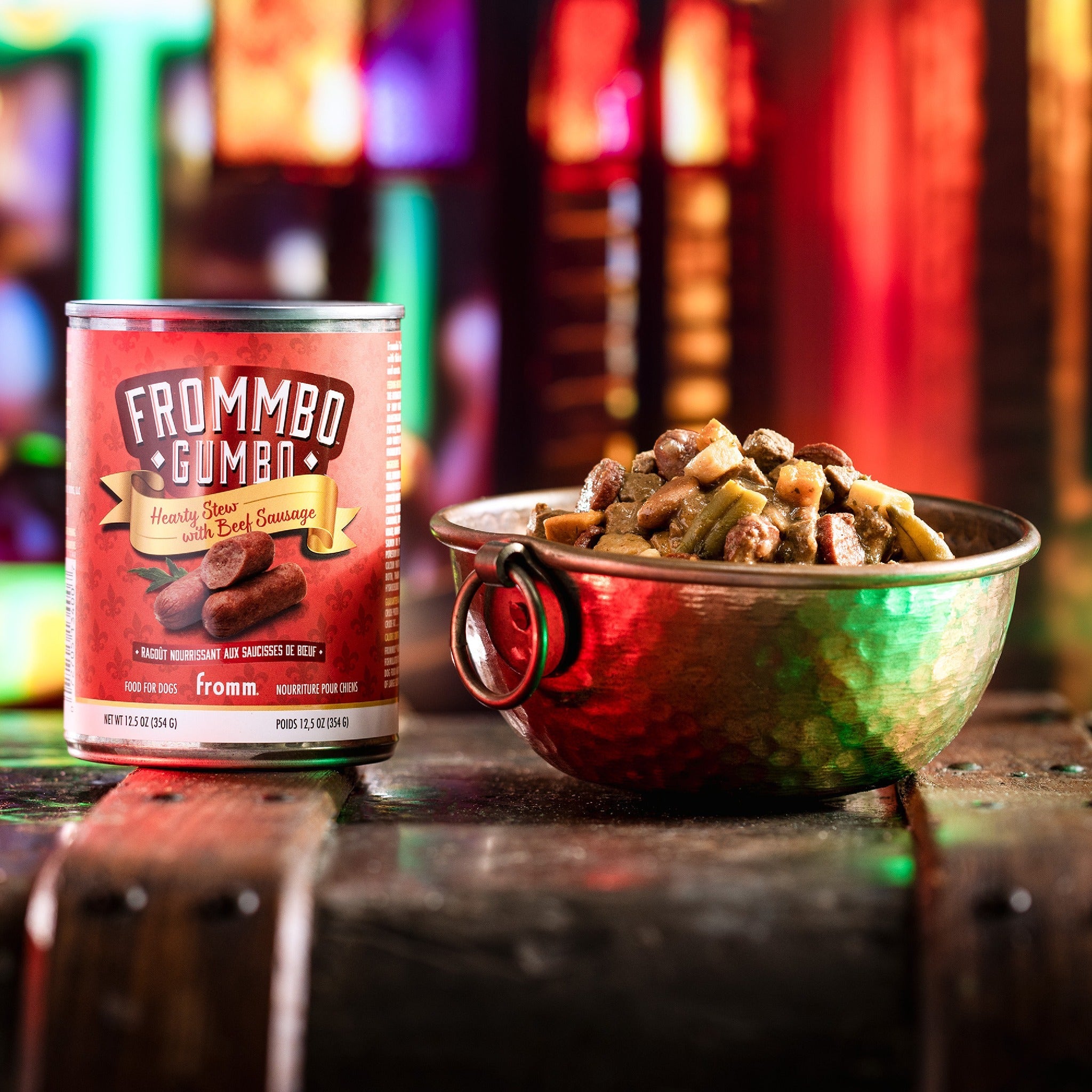 A can of Fromm Frommbo Gumbo Hearty Stew with Beef Sausage beside a filled bowl of stew on a neon-lit night street