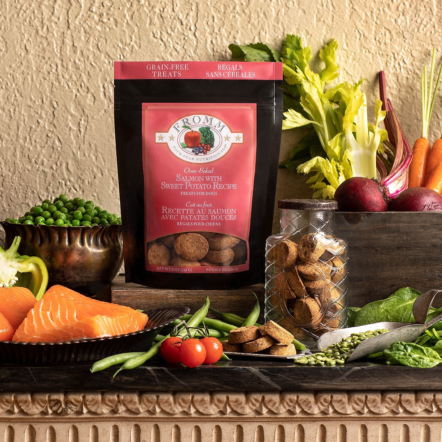 A bag of Fromm Salmon with Sweet Potato Recipe treats among fresh salmon fillets, peas, green beans, carrots, beets, and a jar of oven-baked biscuits