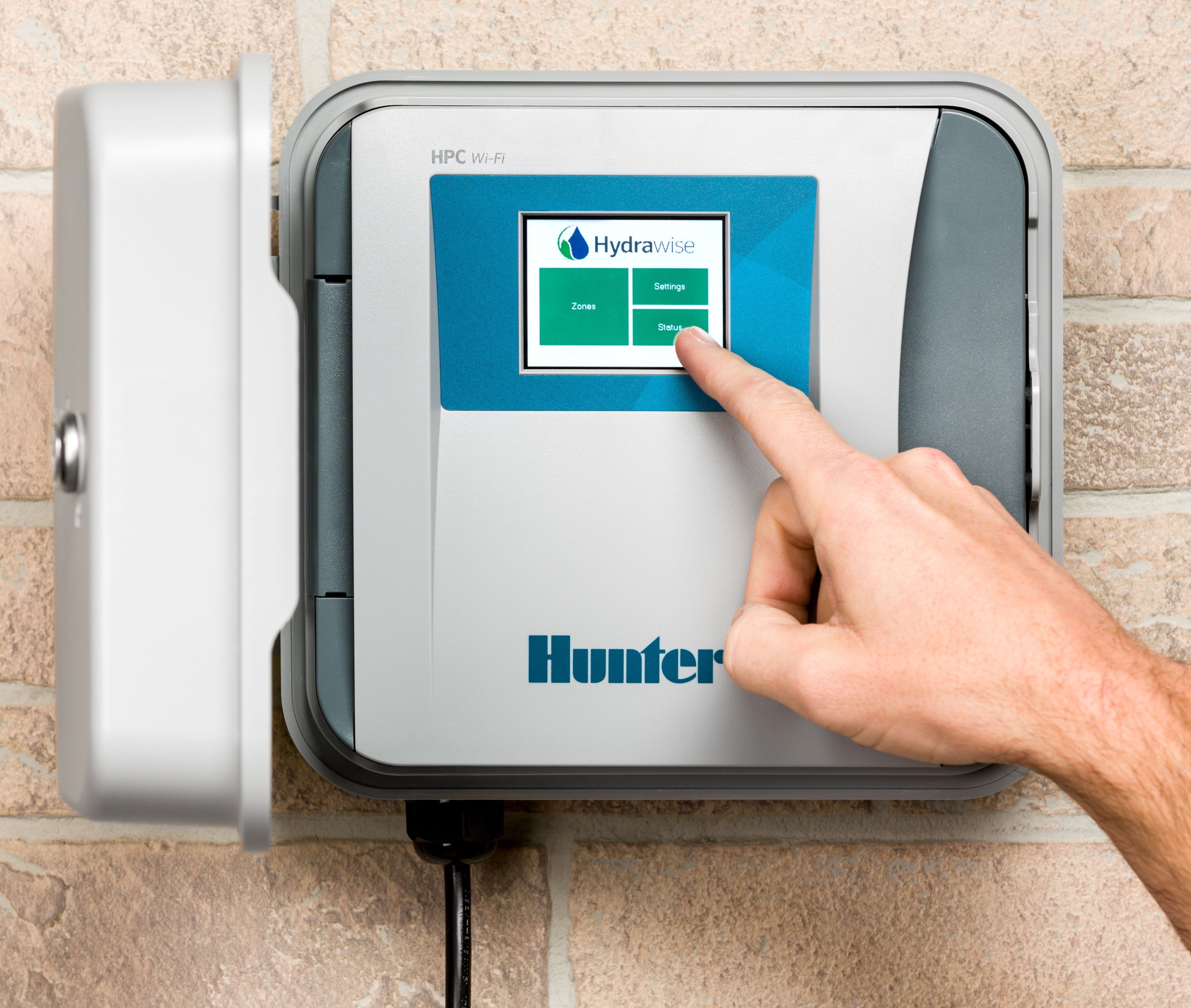 Hunter HPC-400 Hydrawise Wi-Fi controller mounted on an exterior brick wall with a hand using the color touchscreen to access Zones, Settings, and Status tiles