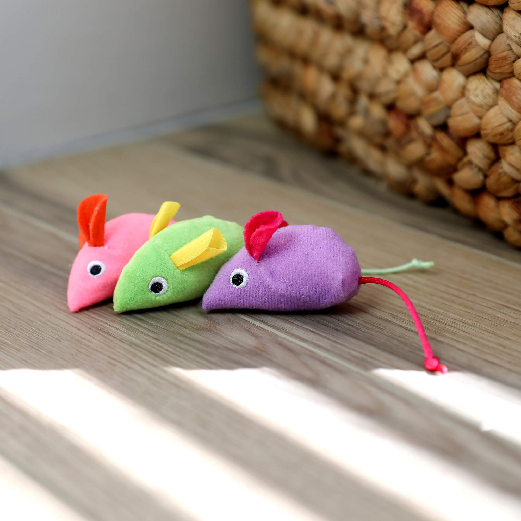 Three Quirky Kitty Glow Mice lined up on a wood floor beside a woven basket
