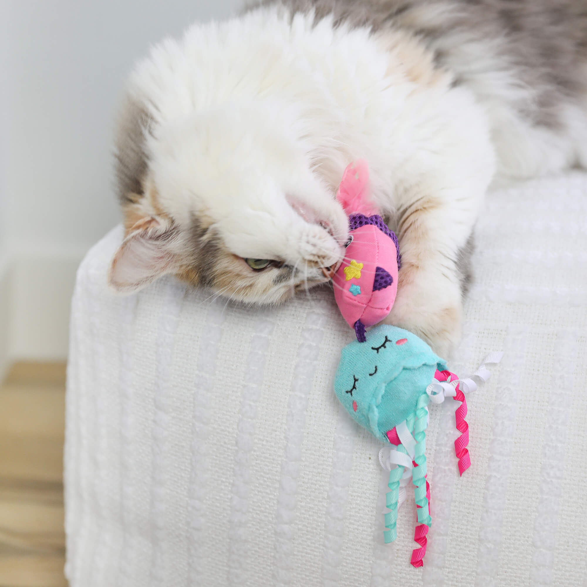 Calico cat lounging on a white sofa cuddling the pink and teal Jellyfish & Chill toys
