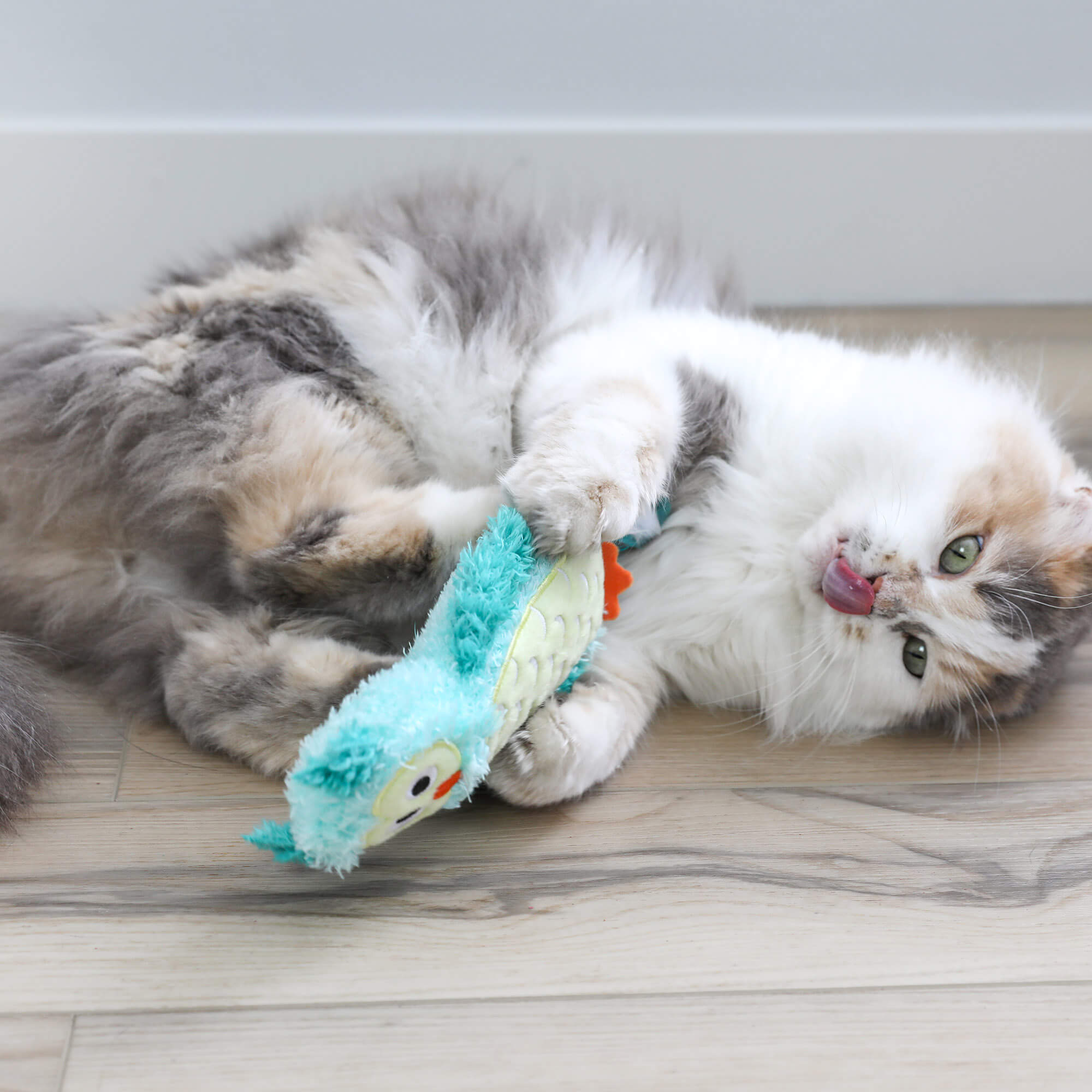 Fluffy calico cat on its back hugging the Night Owl Kicker toy on a wood floor