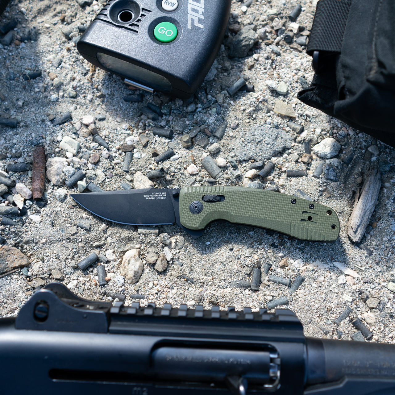 SOG-TAC XR OD Green folding knife on rocky ground next to a shotgun receiver, GPS unit, and spent brass shells in a hunting lifestyle scene