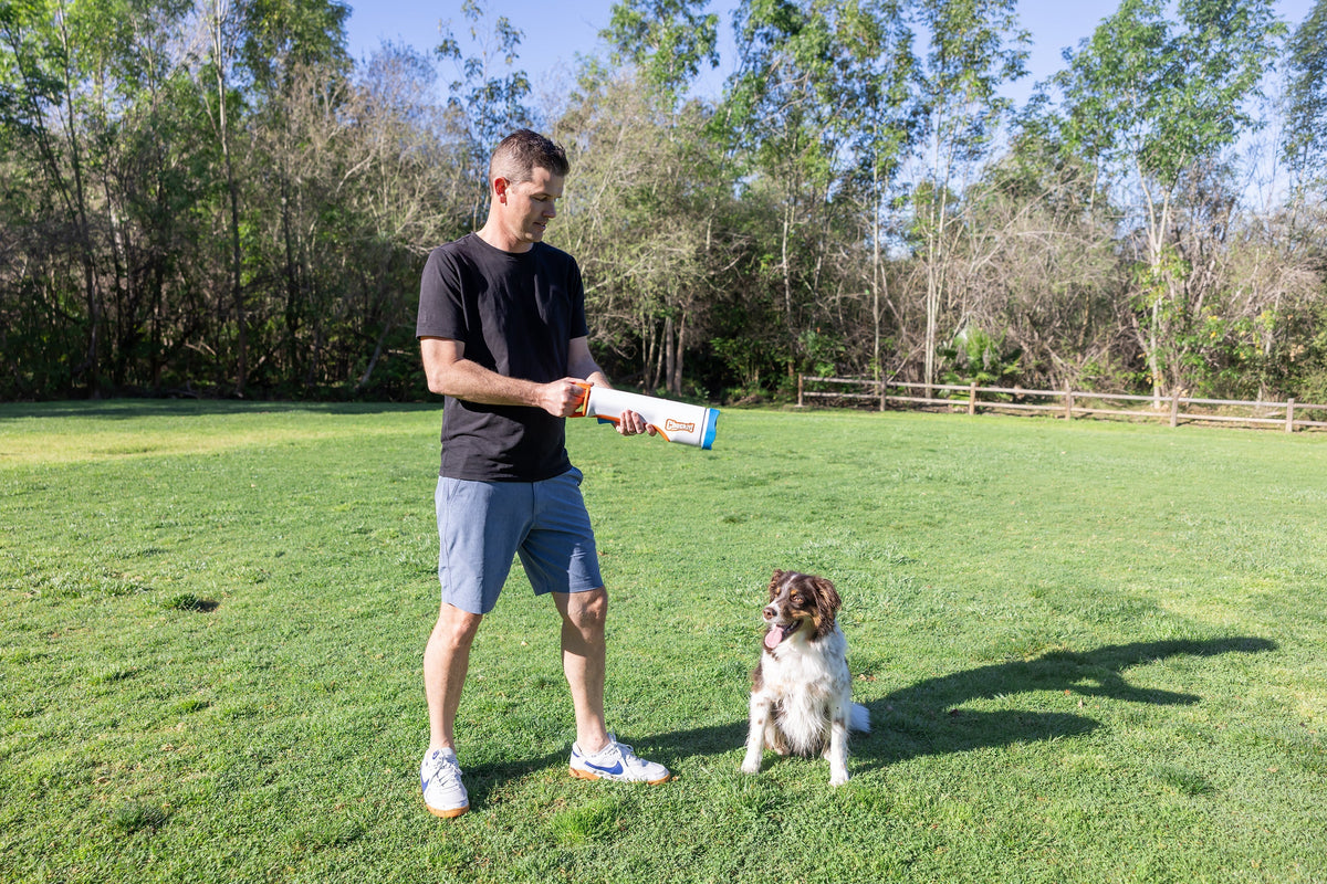 Man in a grassy field ready to launch with the Chuckit! Cannon 16M as his dog waits