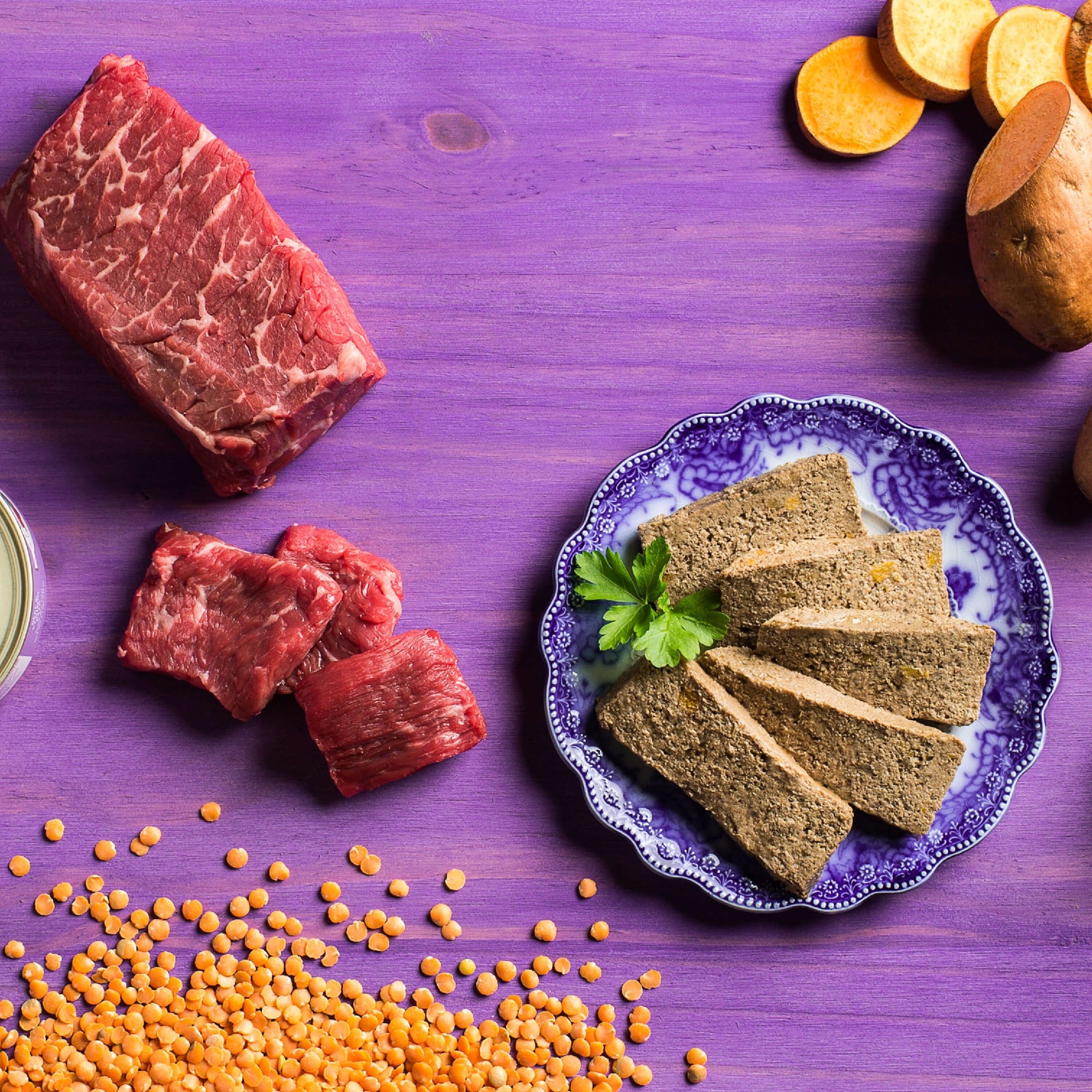 Raw beef cuts, red lentils, and sweet potatoes beside sliced Fromm Beef & Venison Pâté on a blue-patterned plate