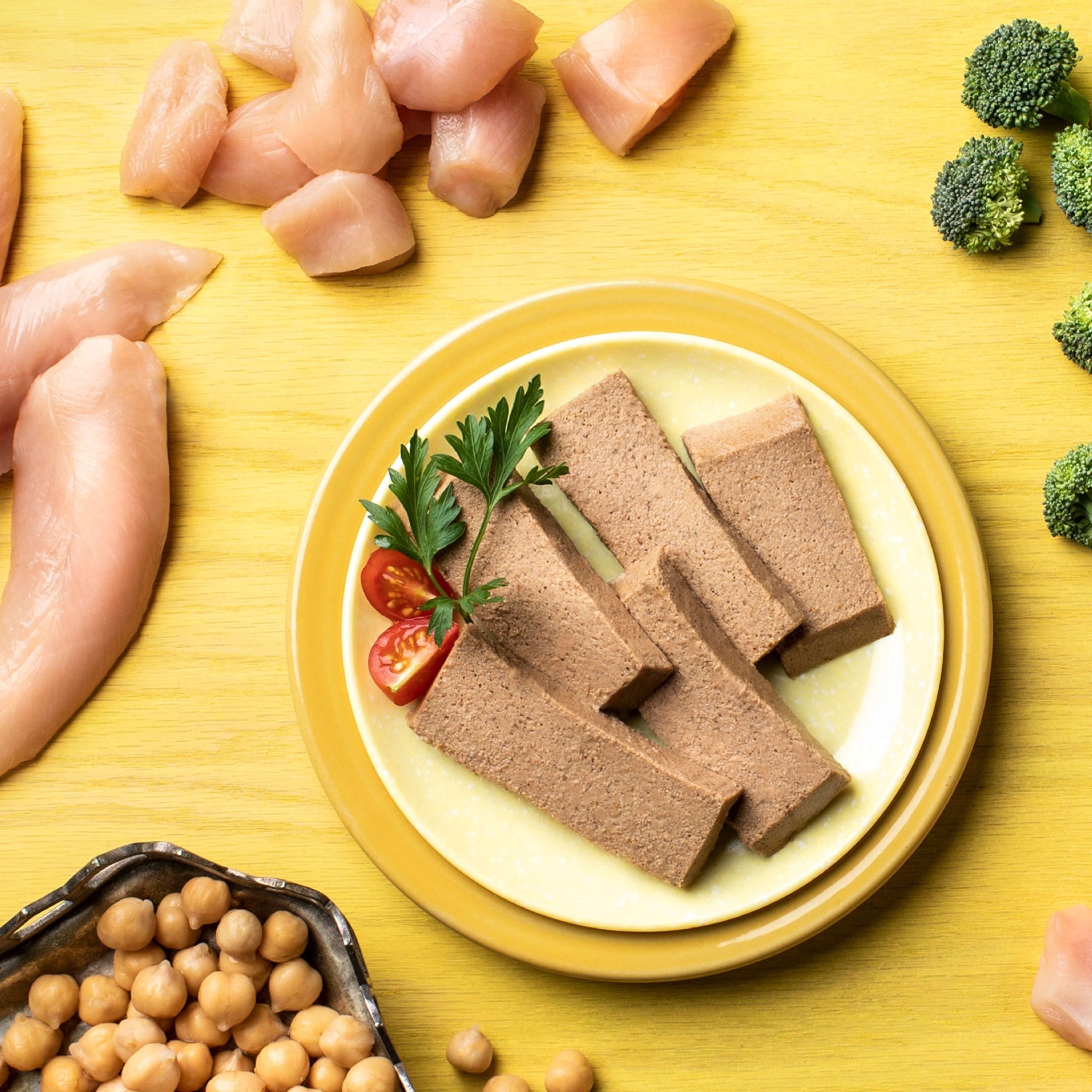 Raw ingredients for Fromm Four-Star Chicken Pâté: raw chicken, chickpeas, broccoli, and sliced pâté on a yellow plate