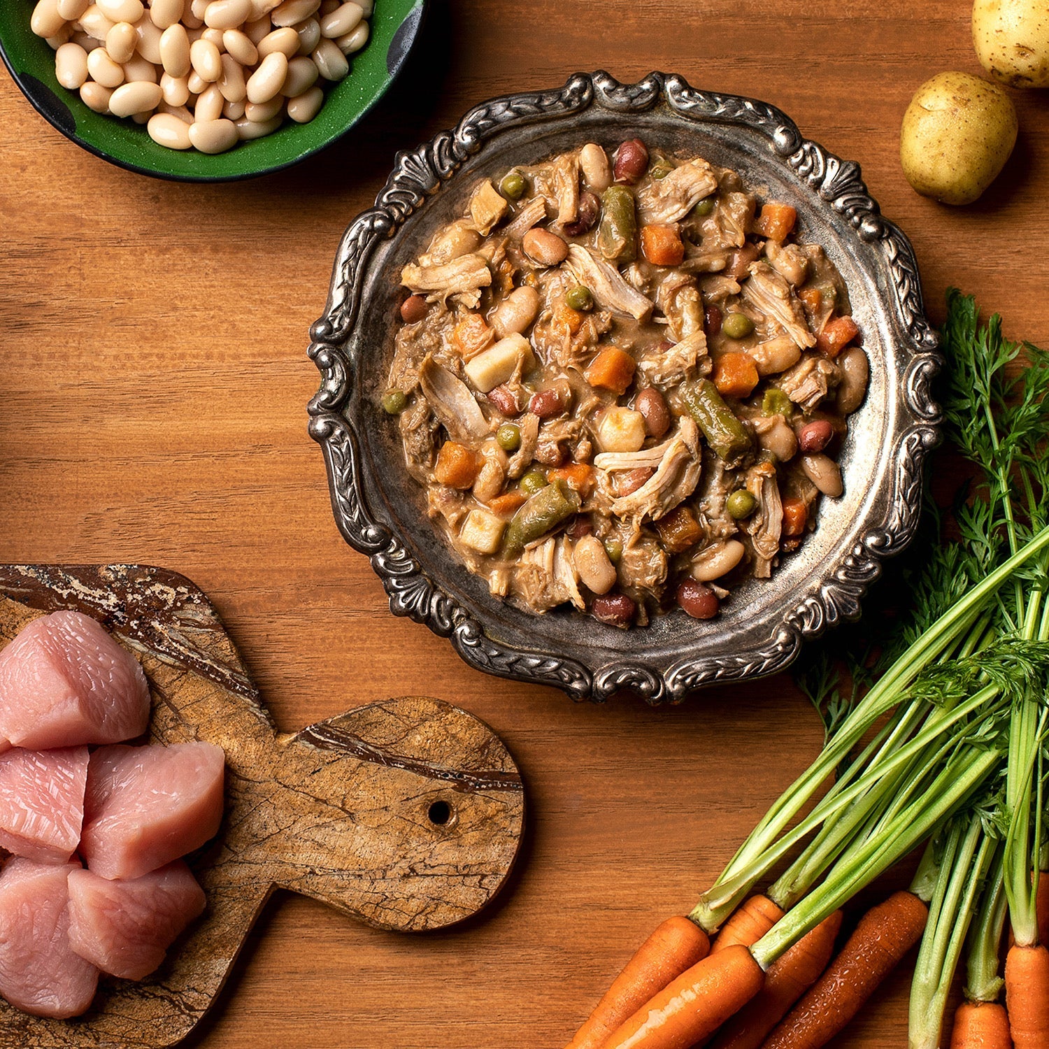 A silver bowl of Fromm Four-Star Shredded Turkey in Gravy Entrée surrounded by raw turkey, white beans, potatoes, and carrots