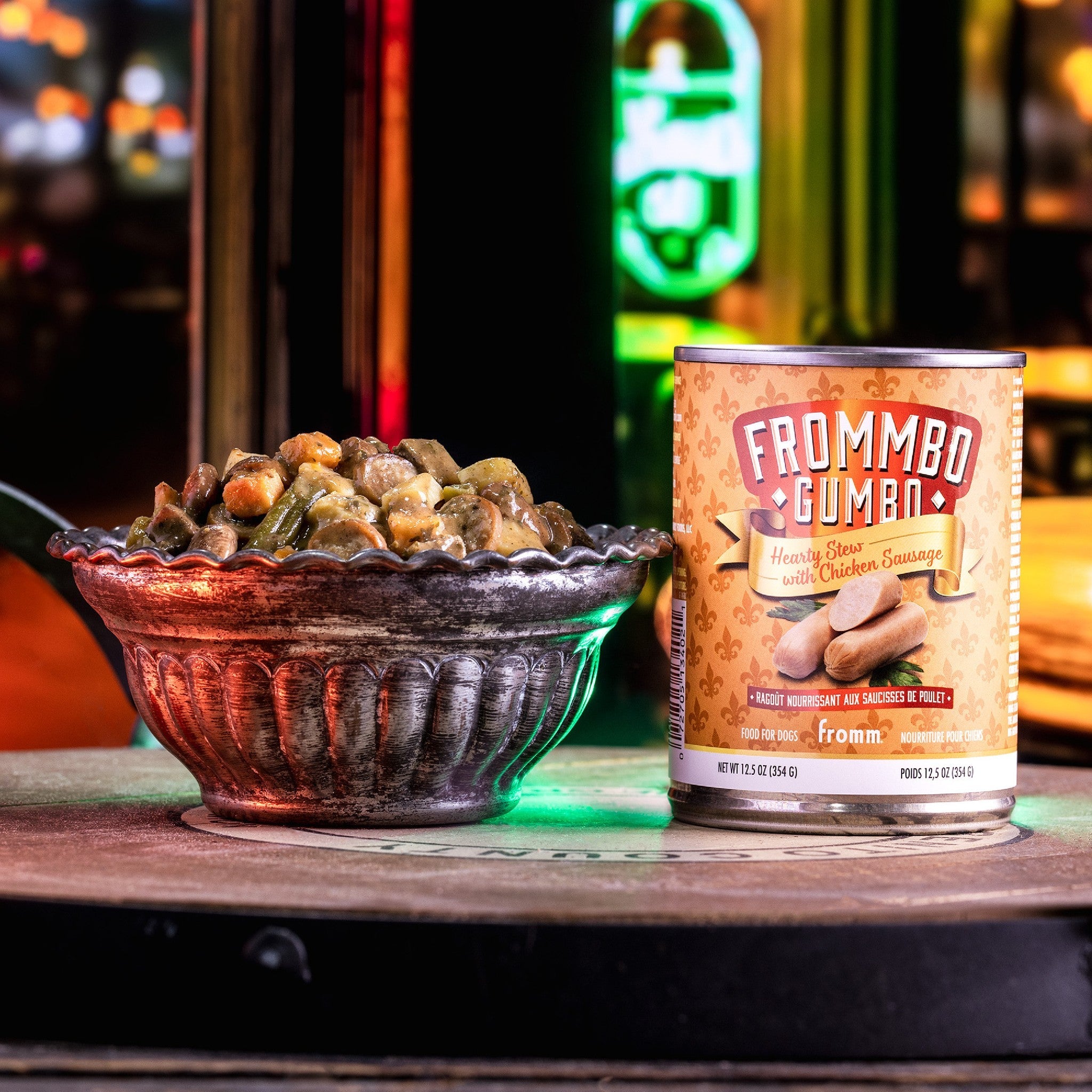 A can of Fromm Frommbo Gumbo Hearty Stew with Chicken Sausage beside a filled bowl of stew on a neon-lit night street