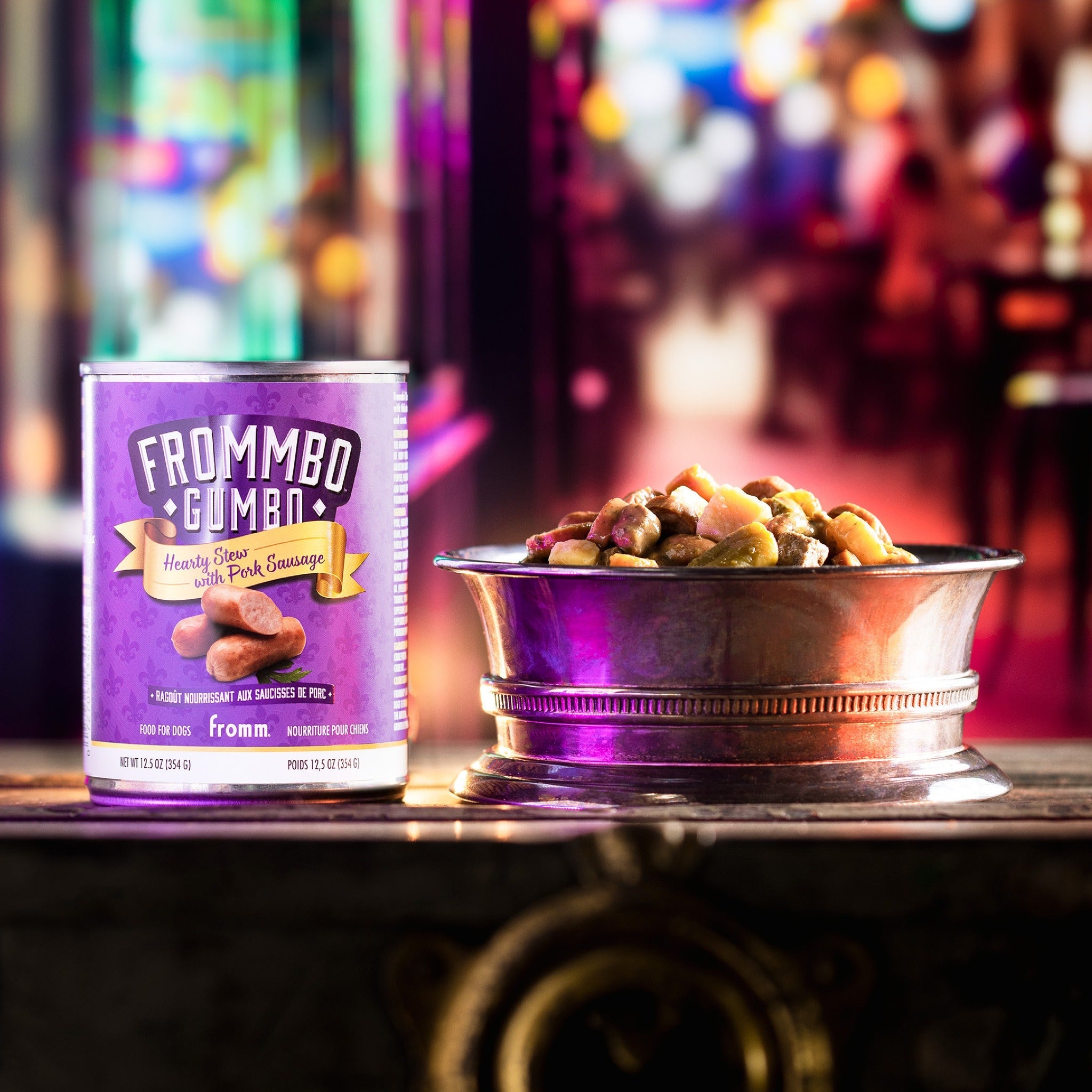 A can of Fromm Frommbo Gumbo Hearty Stew with Pork Sausage beside a filled metal bowl on a neon-lit night street