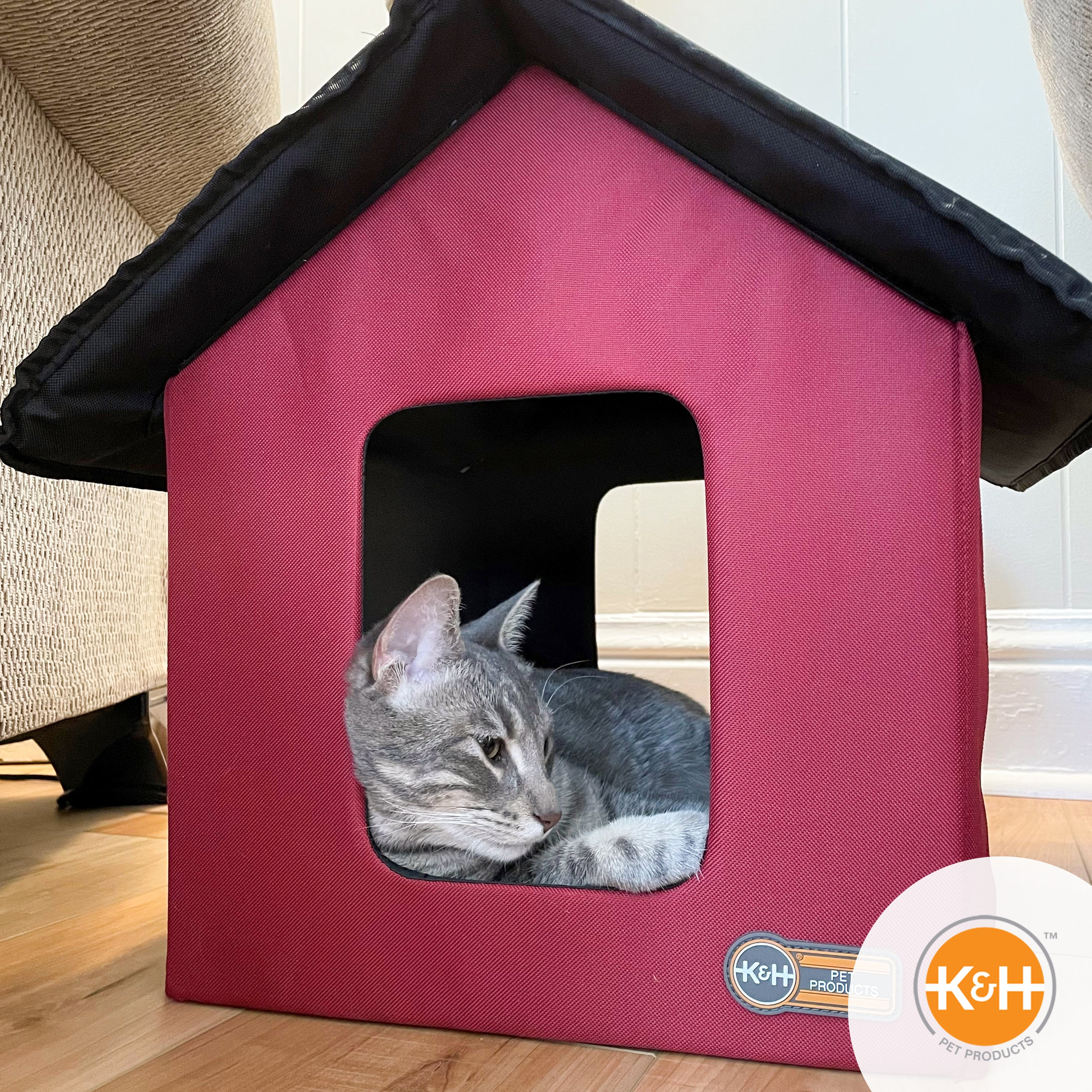 Two cats resting inside a red K&H Outdoor Heated Kitty House placed indoors