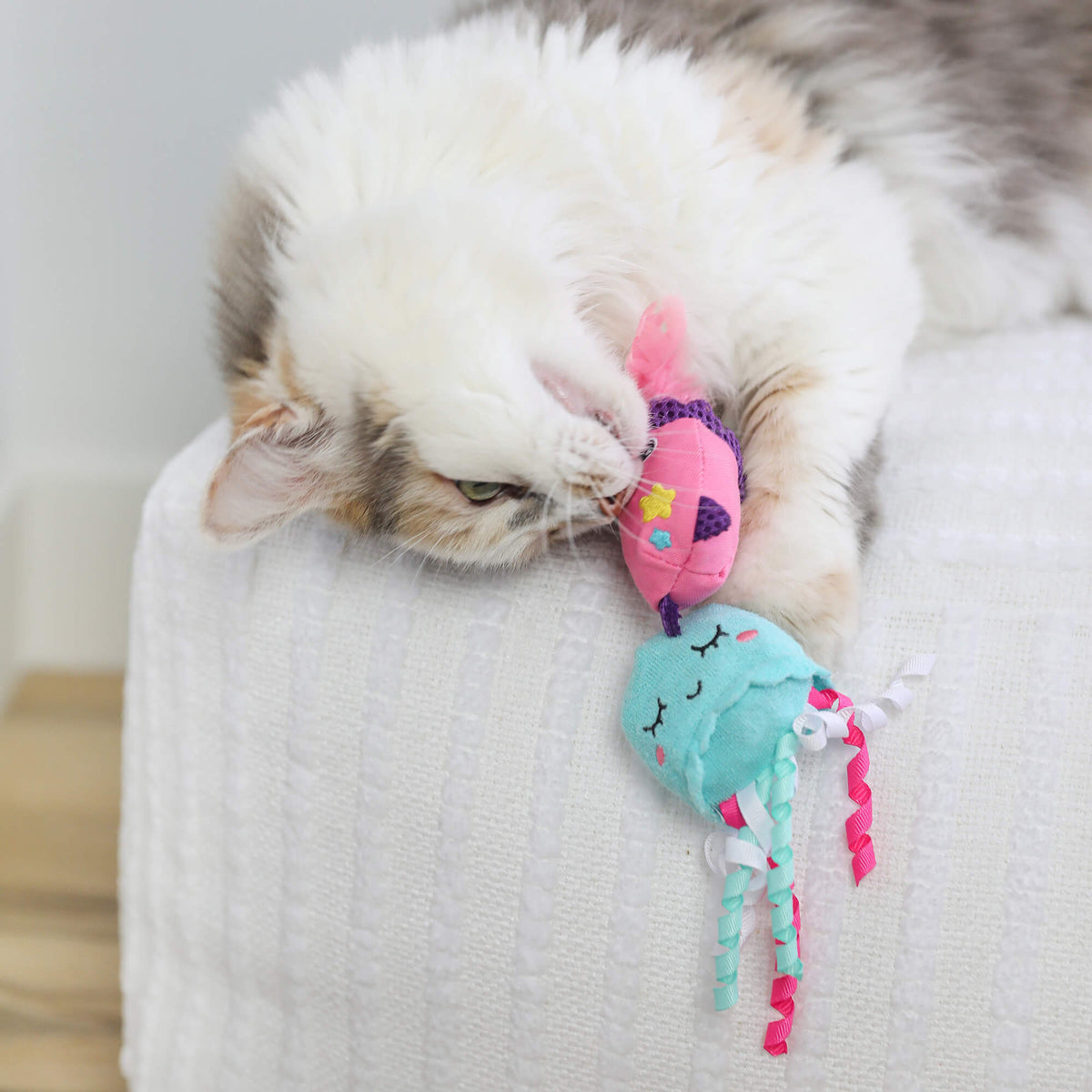 Calico cat lounging on a white sofa cuddling the pink and teal Jellyfish & Chill toys
