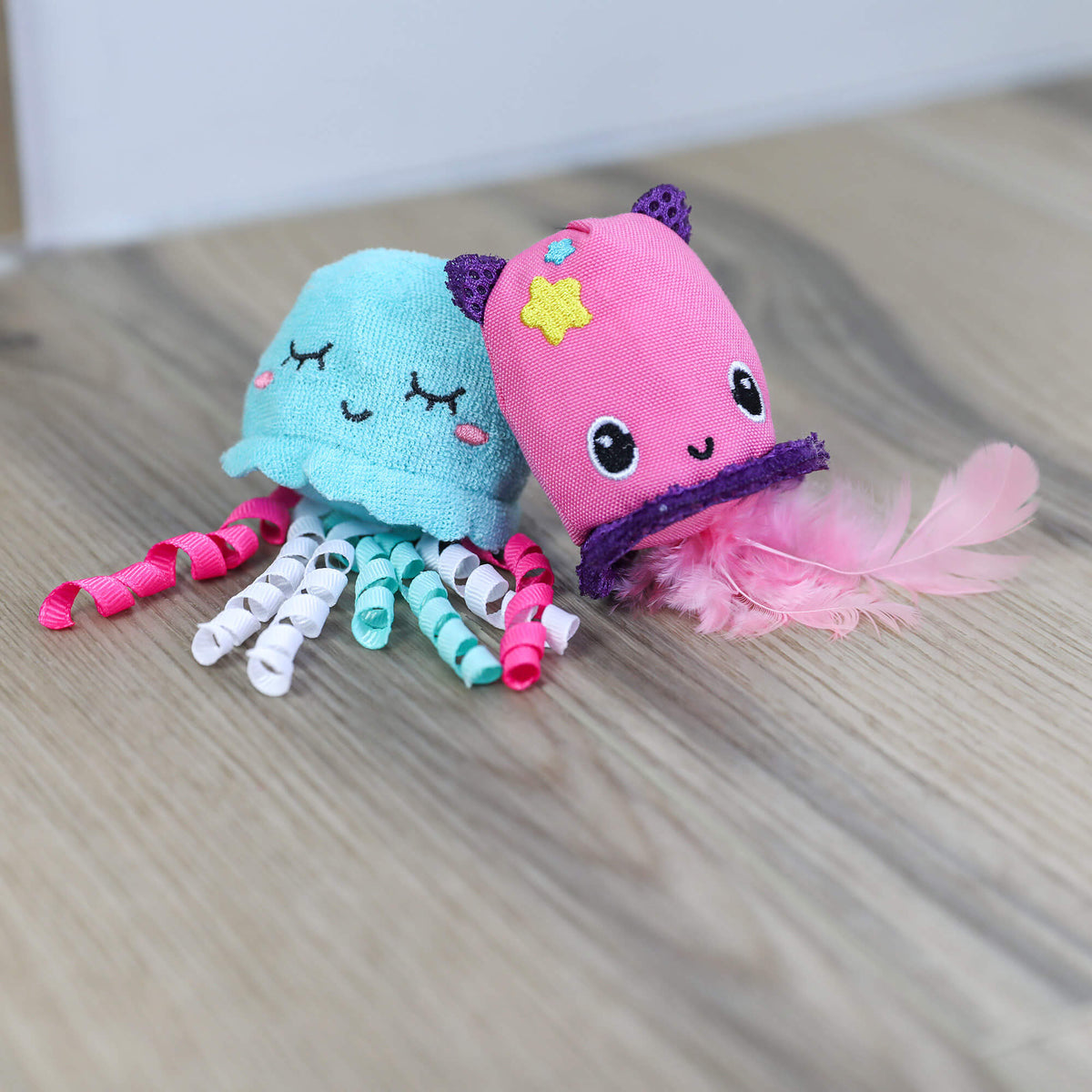 Teal and pink Jellyfish & Chill cat toys sitting side by side on a wood floor