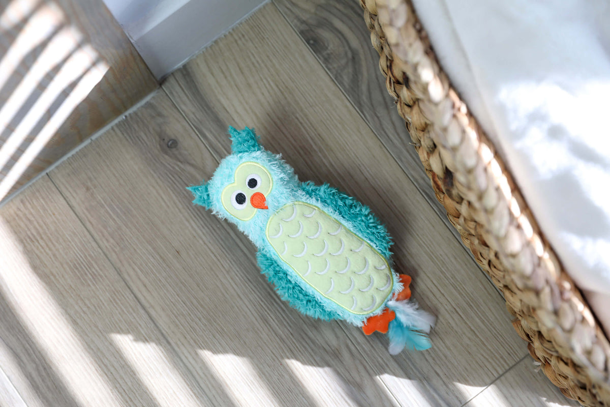 Quirky Kitty Night Owl Kicker plush owl toy lying on a wood floor beside a woven basket