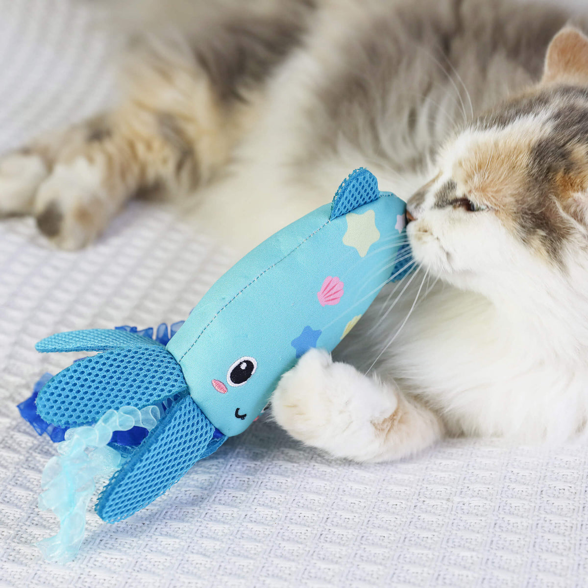 Calico cat sniffing the blue Octopurr Kicker toy while lying on a white blanket