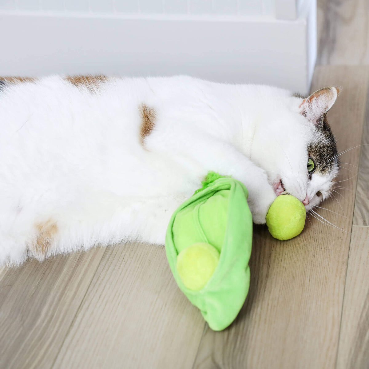 Calico cat lying on the floor chewing a plush pea pulled from the Pea Pawd Kicker pod