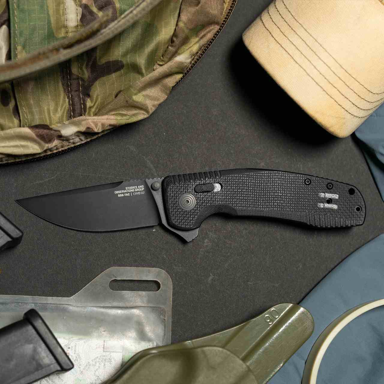 SOG-TAC XR Blackout folding knife on a tactical loadout pad next to multicam jacket holster and field gear