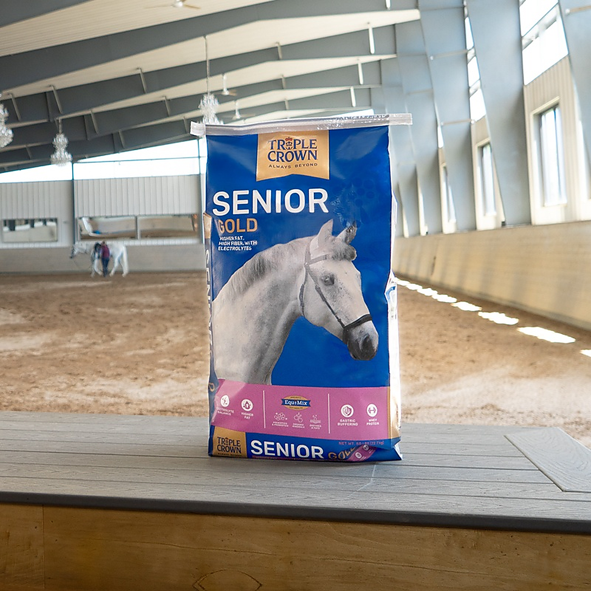 Triple Crown Senior Gold 50 lb bag on an arena ledge with a rider schooling behind it